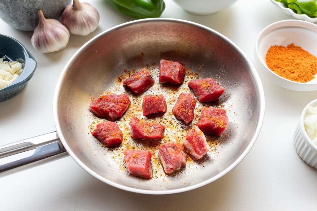 Chunks of raw meat spiced and seared in a stainless steelskillet surrounded by fresh ingredients.