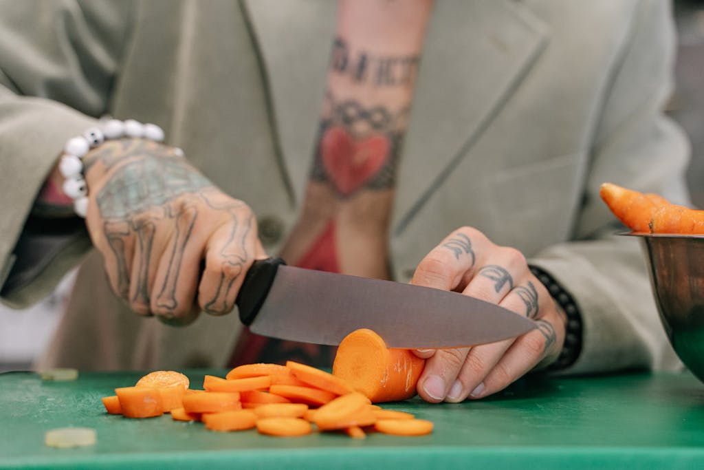 Close-up of tattooed hands slicing carrots on a green board with a chefs knife. Indoors setting.